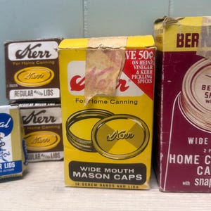 May include: A collection of vintage canning supplies including a blue and white Universal box, two brown Kerr boxes, a yellow Kerr box for wide mouth mason caps, and a tall maroon Bernardin box for wide mouth home canning caps with snap lids.