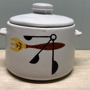 Peut inclure: Un pot en céramique blanc cassé à couvercle et deux petites poignées. Le pot présente un motif fantaisiste d'une fourchette jaune, d'une cuillère marron et d'ustensiles noirs. Le couvercle a un petit bouton pour faciliter le retrait. Un charmant article de cuisine.