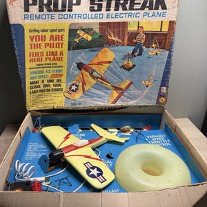 May include: Vintage Prop Streak remote-controlled electric plane toy set. The box features illustrations of the plane in flight. The set includes a yellow and red plane with a star emblem, a control ring, and other accessories.