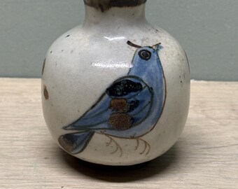 May include: A small, round ceramic bud vase with a speckled off-white glaze. The vase features a hand-painted blue bird illustration with dark brown accents on its wing and tail. The rim of the vase is finished with a dark brown, rustic glaze.
