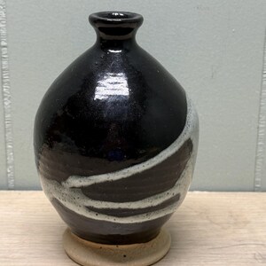 May include: A handmade ceramic vase with a black glazed finish and a white, wavy design. The vase has a rounded body, a narrow neck, and sits on a small, circular base. The pottery is likely for decorative use.