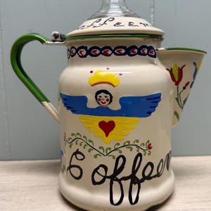 May include: Cream-colored enamel coffee pot with a glass lid and green handle. The pot is decorated with a hand-painted angel, heart, and the word "coffee". Floral and geometric patterns adorn the pot's surface.