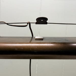 May include: A rectangular, metallic bronze-toned picture light with a horizontal cylindrical housing. It features a thin, arched metal mounting bracket extending above the main body, a small square silver switch, and a black electrical power cord.