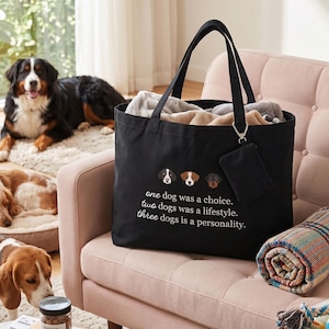 以下が含まれることがあります： 3匹の犬の顔のイラストと「one dog was a choice. two dogs was a lifestyle. three dogs is a personality.」という白い文字がプリントされた黒いトートバッグ。バッグには、シルバーのクリップでハンドルに取り付けられた小さな黒いポーチが付いています。