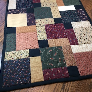 May include: A patchwork quilt table runner with a black border. The runner is made of various fabrics in shades of brown, green, red, and white. The fabrics feature floral and geometric patterns.