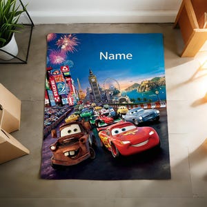 May include: A rectangular blanket featuring a colorful graphic print of characters from the movie Cars, including a red race car and a brown tow truck, set against a city skyline with fireworks and the word Name printed in white at the top.