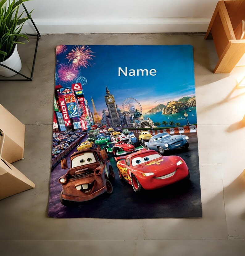 May include: A rectangular blanket featuring a colorful graphic print of characters from the movie Cars, including a red race car and a brown tow truck, set against a city skyline with fireworks and the word Name printed in white at the top.