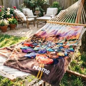 May include: A soft, rectangular blanket featuring a colorful graphic print of various animated race cars from the movie Cars, set against a desert landscape. The blanket includes the word Name printed in white text near the bottom edge.