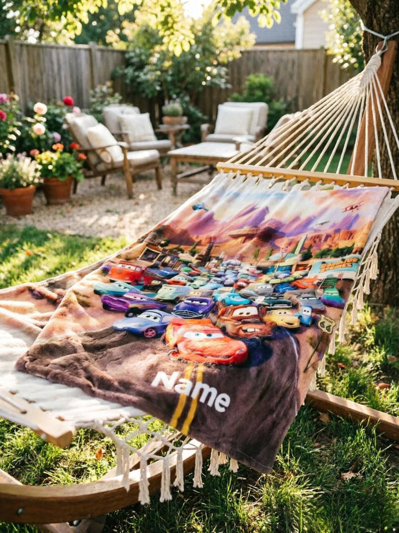 May include: A soft, rectangular blanket featuring a colorful graphic print of various animated race cars from the movie Cars, set against a desert landscape. The blanket includes the word Name printed in white text near the bottom edge.