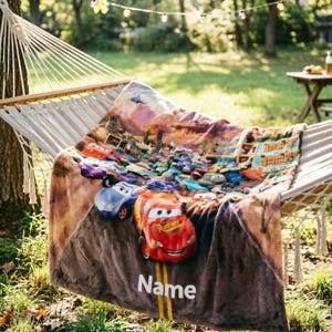 May include: A soft plush throw blanket featuring a colorful graphic print of various animated race cars from the movie Cars, including a prominent red race car in the foreground, with the word Name printed in white text on a road-like design.