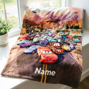 May include: A soft plush blanket featuring a colorful graphic print of the Radiator Springs town scene from the movie Cars, with Lightning McQueen in the foreground and the word Name printed in white at the bottom of the road design.