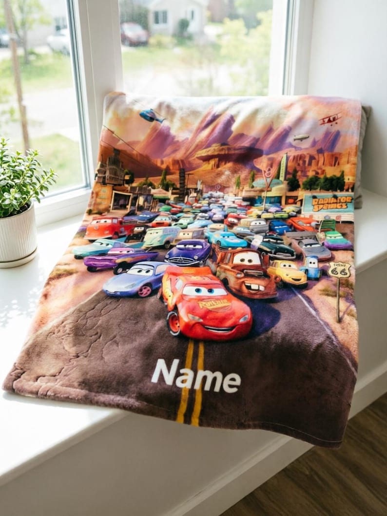 May include: A soft plush blanket featuring a colorful graphic print of the Radiator Springs town scene from the movie Cars, with Lightning McQueen in the foreground and the word Name printed in white at the bottom of the road design.