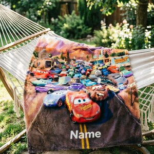 May include: A soft fleece blanket featuring a colorful graphic print of various animated race cars from the movie Cars, including Lightning McQueen and Mater, driving on a road. The word Name is printed in white text at the bottom of the blanket.