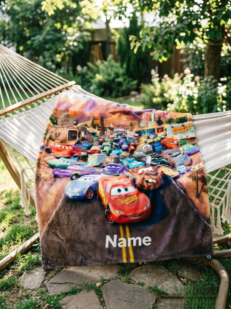 May include: A soft fleece blanket featuring a colorful graphic print of various animated race cars from the movie Cars, including Lightning McQueen and Mater, driving on a road. The word Name is printed in white text at the bottom of the blanket.