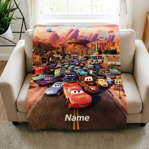 May include: A soft throw blanket featuring a vibrant scene from the movie Cars, with Lightning McQueen in the foreground on a desert road. The background shows Radiator Springs with red rock formations and the word Name printed at the bottom.
