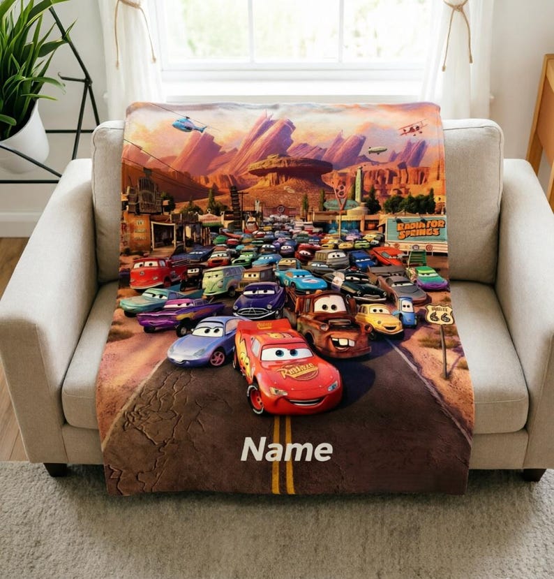 May include: A soft throw blanket featuring a vibrant scene from the movie Cars, with Lightning McQueen in the foreground on a desert road. The background shows Radiator Springs with red rock formations and the word Name printed at the bottom.