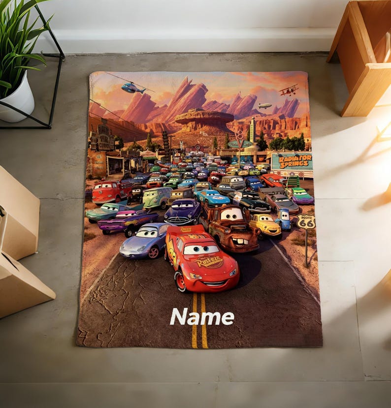 May include: A rectangular blanket featuring a colorful graphic print of the animated movie Cars, showcasing Lightning McQueen and various other vehicle characters on a road in Radiator Springs, with the word Name printed in white at the bottom.