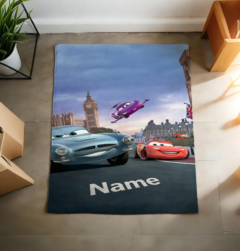 May include: A rectangular blanket featuring a scene from the movie Cars 2 with Lightning McQueen, Finn McMissile, and Holley Shiftwell driving in London near Big Ben. The word Name is printed in white capital letters on the dark road surface.