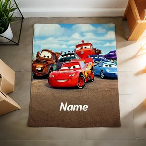 May include: A rectangular fleece blanket featuring a colorful graphic of various animated car characters, including a red race car with the number 95, a brown tow truck, and a blue sports car, with the word Name printed in white at the bottom.