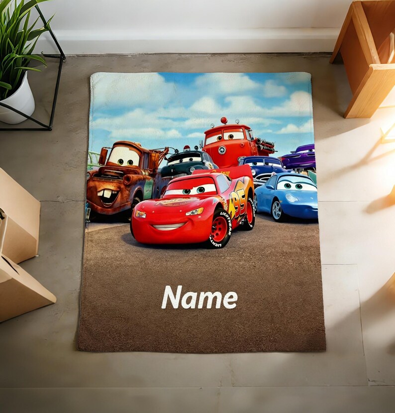 May include: A rectangular fleece blanket featuring a colorful graphic of various animated car characters, including a red race car with the number 95, a brown tow truck, and a blue sports car, with the word Name printed in white at the bottom.