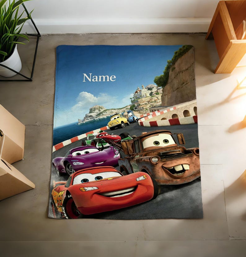 May include: A rectangular fleece blanket featuring a colorful graphic print of characters from the movie Cars, including Lightning McQueen and Mater, racing on a coastal track. The word Name is printed in white text at the top of the blanket.