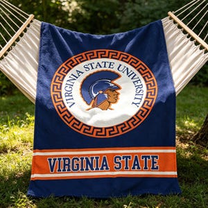 May include: A navy blue fleece blanket featuring the Virginia State University logo in the centre, which includes a Trojan mascot graphic inside an orange and white circular border. The text Virginia State is printed in blue on an orange stripe.