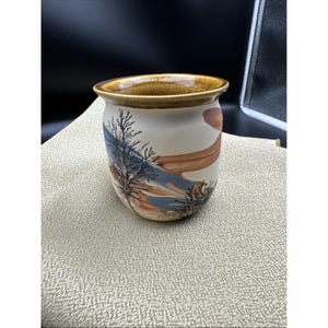 May include: A ceramic mug with a rounded body and a flared rim. The exterior features a cream-colored base with painted landscape elements including dark tree silhouettes, muted blue shapes, and soft orange streaks. The interior rim is glazed in brown.