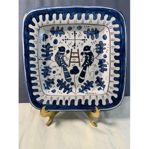 May include: Square ceramic serving dish with a white background and blue border. The dish features a hand-painted design of two birds, a ladder, and floral elements in blue and brown. The dish is supported by a gold-colored stand.