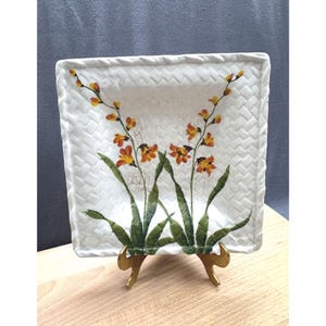 Puede incluir: Plato cuadrado de cerámica blanca con borde de textura tejida. El plato presenta un diseño floral con flores naranjas y amarillas y hojas verdes. El plato se exhibe sobre un soporte de metal dorado.