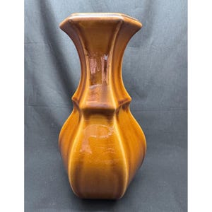 May include: A ceramic vase with a glossy, multi-tonal brown glaze. The vessel features a wide, rounded base that tapers into a narrow neck, which then flares outward at the top rim. The surface displays vertical ridges and a smooth, reflective finish.