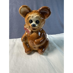 Può includere: Figurina vintage a forma di orso in ceramica con finitura marrone lucida. L'orso ha grandi occhi neri, un piccolo fiocco rosa e un'espressione sorridente. La statuina è seduta, con le braccia e le gambe leggermente curve.