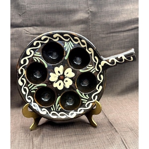 May include: A black ceramic escargot dish with seven round indentations. The dish features a white and green floral design around the rim and a central flower. It has a handle and is displayed on a gold-colored stand.