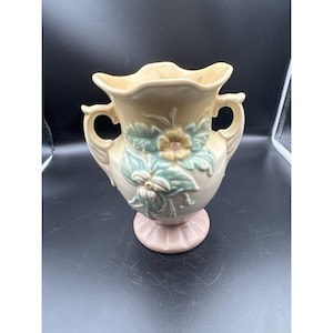 Puede incluir: Un jarrón de cerámica con cuerpo de color crema, que presenta dos asas laterales y un borde festoneado. La parte frontal está decorada con motivos florales en relieve en tonos verde pálido y amarillo, sobre una base de pedestal estriada.