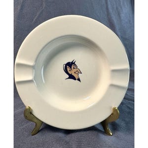 May include: White ceramic ashtray with a blue and tan devil logo in the center. The ashtray has three rests for cigarettes and is displayed on a gold-colored metal stand. The ashtray is round and has a slightly raised edge.