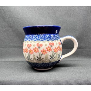 Op de afbeelding: Een keramische mok met een bolle vorm, voorzien van een donkerblauwe rand en voet. Het lichaam is versierd met een herhalend patroon van oranje-rode bloemen met groene stelen en bladeren, geaccentueerd door kleine blauwe cirkelvormige motieven op een crèmekleurige achtergrond.