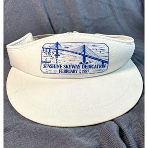 May include: White visor featuring a blue bridge graphic and the text "Sunshine Skyway Dedication February 7, 1987." The visor has a curved peak and a padded headband.