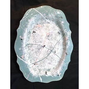 May include: An oval-shaped serving platter with a scalloped edge, featuring a light teal base colour decorated with a white speckled paint splatter pattern, thin intersecting lines, and small scattered pink and dark grey geometric accents.