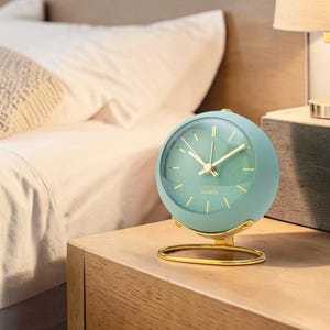May include: A mint green quartz alarm clock with gold-colored hands and a gold-colored base. The clock is round and sits on a wooden nightstand. The word "QUARTZ" is printed on the clock face.