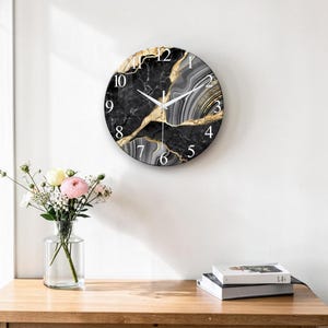 May include: Round wall clock with a black marble design and gold accents. The clock face features white numerals and hands. The clock is hanging on a white wall. A vase of flowers and books are on a wooden table.