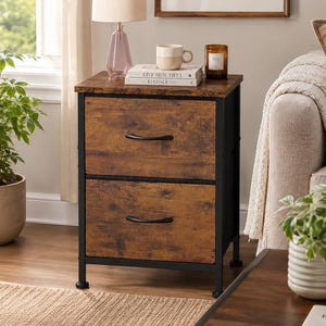 May include: A two-drawer nightstand with a rustic brown wood-grain finish and a black metal frame. The unit features two drawers with curved black handles, topped with a lamp, a ceramic mug, and a book titled Live Beautiful by Athena Calderone.