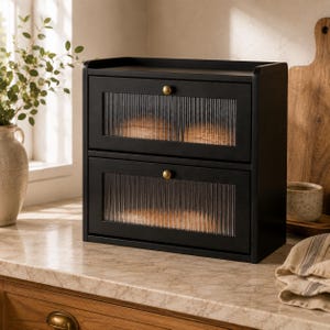 May include: A two-tier black bread box featuring fluted glass panels on the front of each drop-down door. Each door is equipped with a small, round, brass-colored knob. The structure is rectangular with a flat top surface for additional storage.