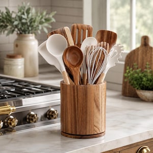 May include: A cylindrical wooden utensil holder containing a collection of kitchen tools. The set includes various wooden spoons, spatulas, and slotted turners, alongside white silicone-tipped ladles, whisks, and a pasta server with wooden handles.