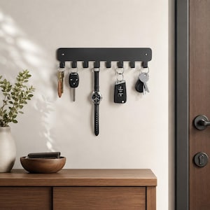 May include: A matte black wall-mounted organizer rack featuring ten evenly spaced hooks. The rack is currently holding various items including a set of keys, a car key fob, and a wristwatch with a black leather strap, all suspended from the hooks.