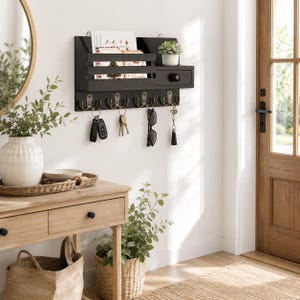 May include: A black wall-mounted entryway organizer featuring a mail slot, a small storage drawer with a round knob, and four double metal hooks. The organizer holds various envelopes, a small potted succulent, keys, and sunglasses.