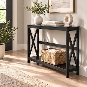 May include: A black console table featuring an X-shaped design on both side panels. The table has a flat top surface and a lower shelf. A woven basket sits on the bottom shelf, while a white vase and two books are placed on the top.