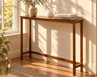 May include: A tall, narrow wooden console table with a rectangular top and four straight legs. On the surface sits a textured ceramic vase containing green leafy branches and two stacked books titled The Kinfolk and At Home.