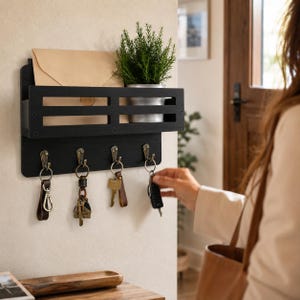 May include: A black wooden wall-mounted key holder with four hooks, a shelf, and a slot for mail. The key holder has a small potted plant and a brown envelope on the shelf. Keys hang from the hooks.
