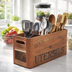 May include: A wooden utensil holder with the words "COOK EAT BAKE UTENSILS" in black lettering. The holder contains various kitchen utensils, including spatulas, spoons, and serving spoons. A blender and a bowl of fruit are in the background.