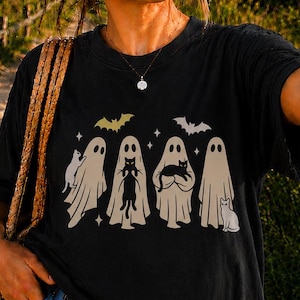 Pu&ograve; includere: Una t-shirt nera con una stampa grafica di quattro fantasmi beige, ognuno che interagisce con un gatto nero, accentuata da due piccoli pipistrelli e stelle. Indossata con pantaloncini di jeans blu e una borsa di paglia intrecciata.