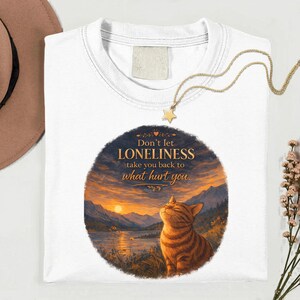 May include: White t-shirt featuring a round graphic with a cat and a sunset landscape. The graphic includes the text "Don't let loneliness take you back to what hurt you." A gold necklace with a star pendant is draped on the shirt.